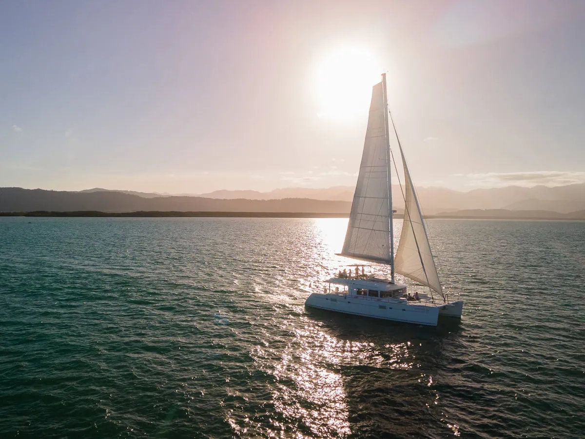 a large sailing catamaran sailing on the ocean during sunset with mountains in the background 